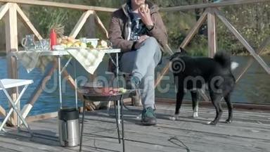 成熟的男人，穿着温暖的衣服，坐在桌子旁，吃着汉堡，靠近一只饥饿的狗，在河岸上野餐
